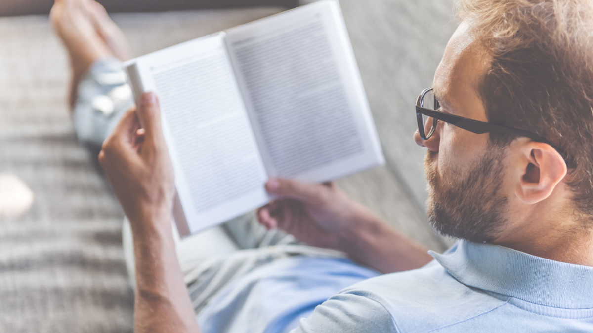 Back view of handsome young businessman in casual clothes and eyeglasses reading a book while lying on couch at home.