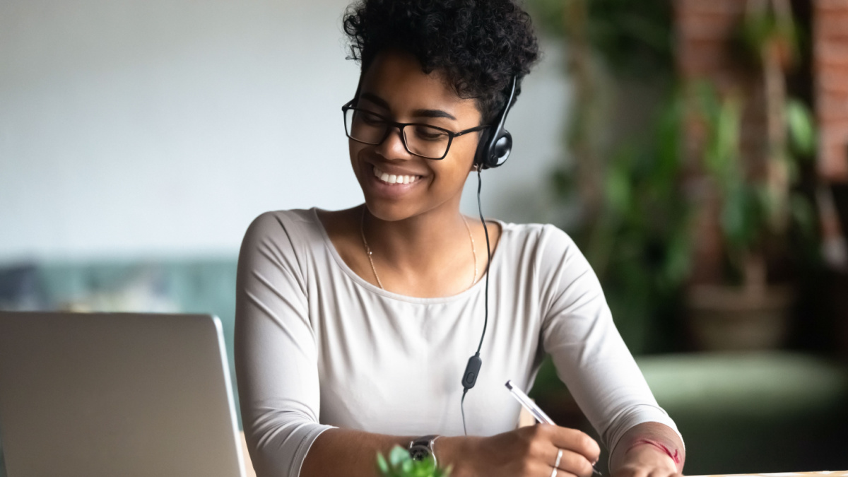 Smiling african American millennial female student in headphones and glasses sit at desk watch webinar making notes.
