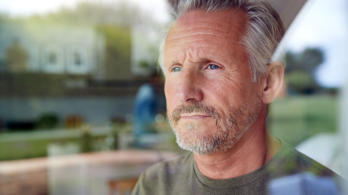 Concerned Senior Man Standing And Looking Out Of Kitchen Door Viewed Through Window.