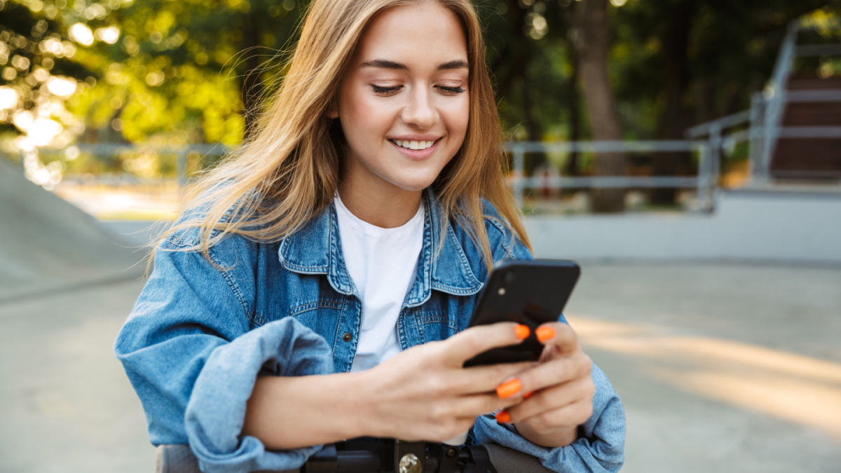 Photo of a positive cheerful teenage girl in park walking on scooter using mobile phone.