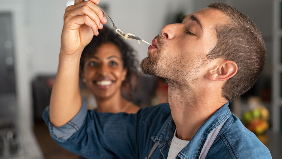 Man tasting spaghetti pasta while smiling woman look at him. Closeup face of young man tasting meal while cooking at home. Handsome guy eating noodles with fork in kitchen and feel his recipe.