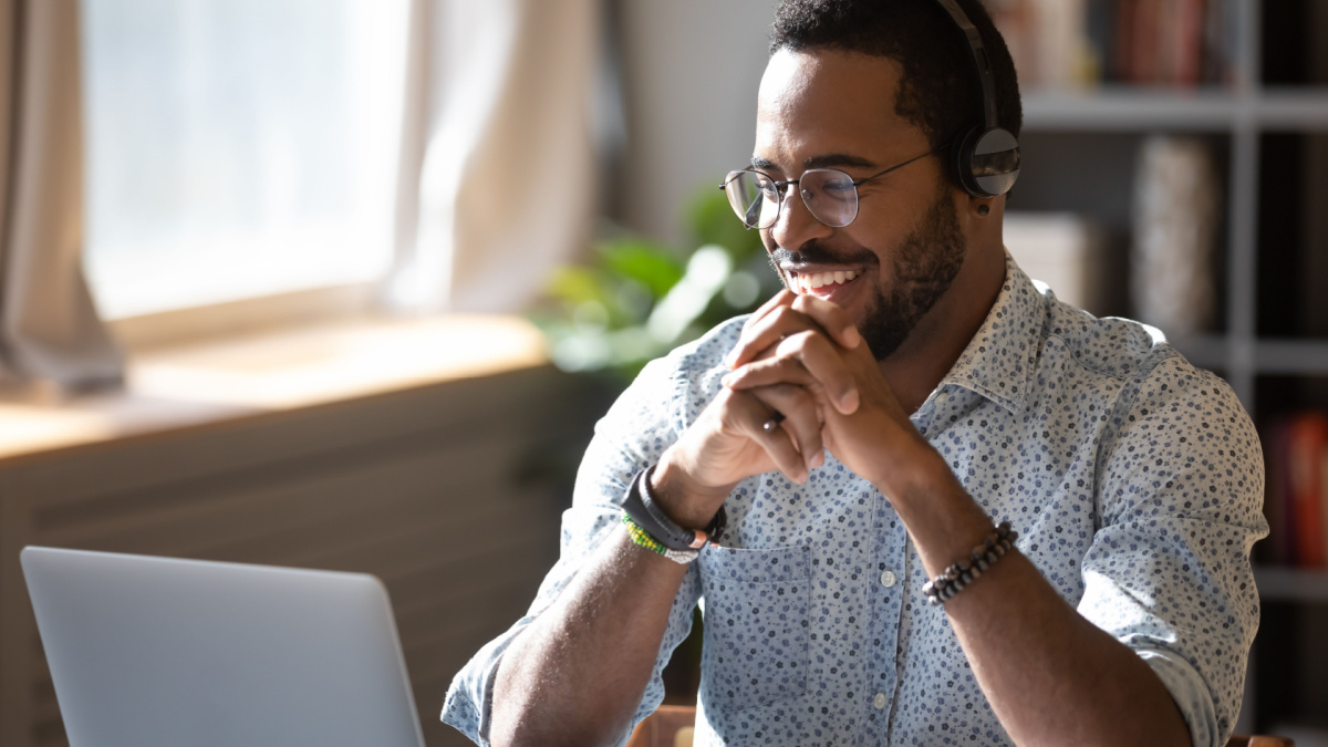 Happy millennial african american man in glasses wearing headphones, enjoying watching educational webinar on laptop.