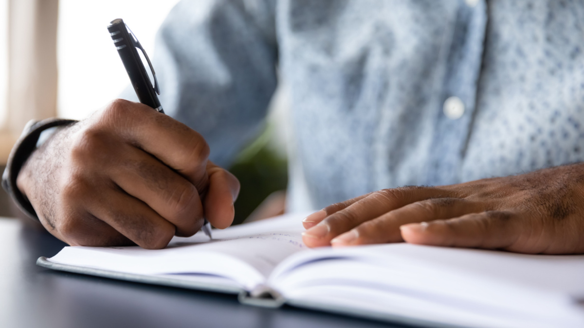 Close up of african American male student busy handwriting in notebook preparing for exam or test, biracial man worker make notes or list writing down, planning or thinking at workplace in office.