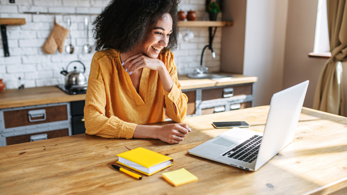 A cheerful mixed-race girl uses laptop for remote work or home leisure while sitting in the kitchen at home.