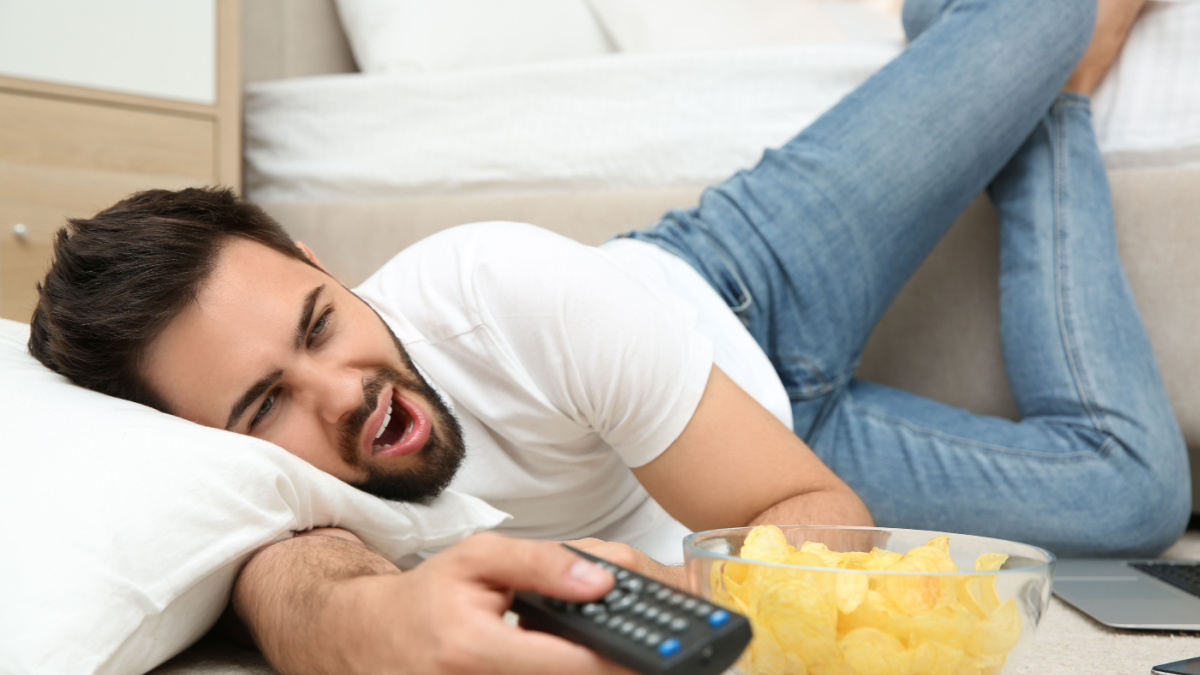 Lazy young man with bowl of chips watching TV while lying on floor at home.