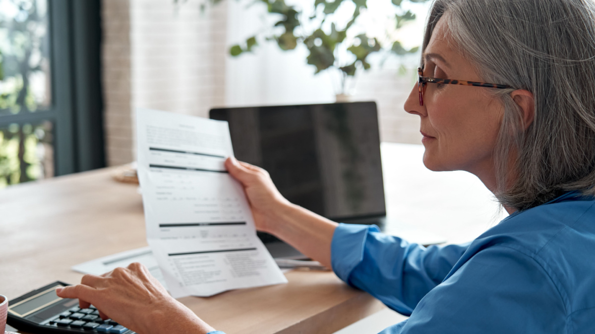 Senior mature business woman holding paper bill using calculator, old lady managing account finances, calculating money budget tax, planning banking loan debt pension payment sit at home office table.