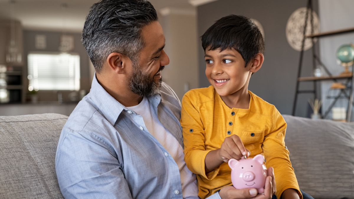 Indian father and smiling son putting coin into piggy bank.