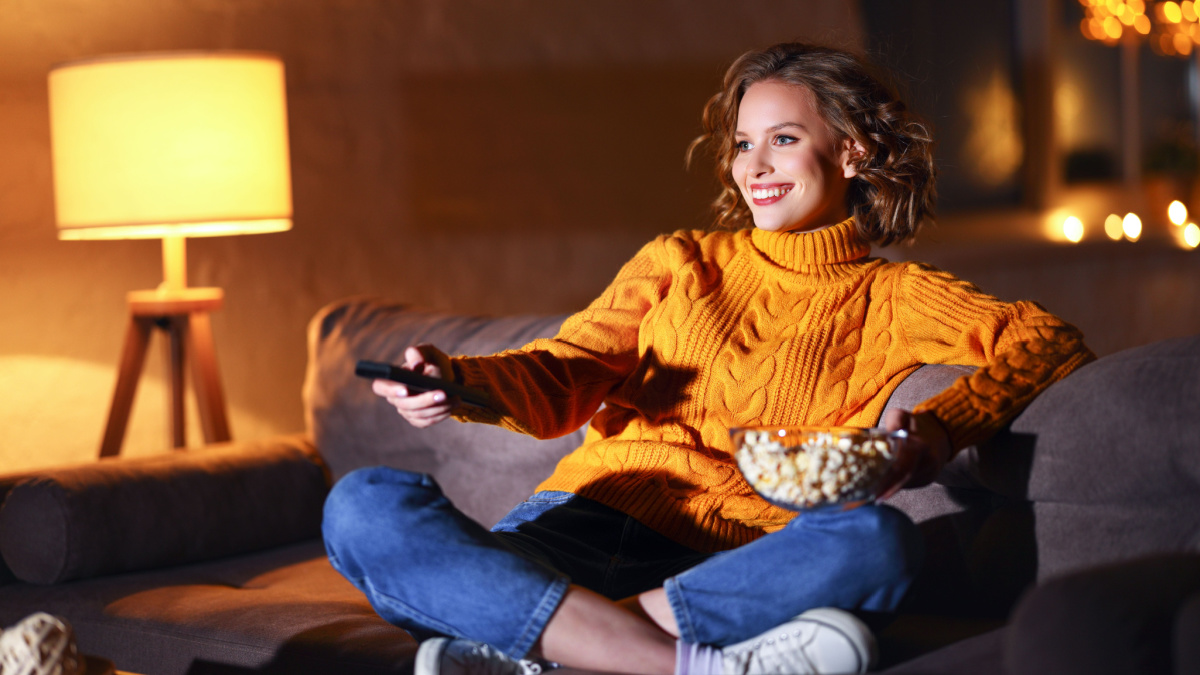young cheerful woman eating popcorn and watches movie on cable TV while switching channels with the remote control at home in evening alone.