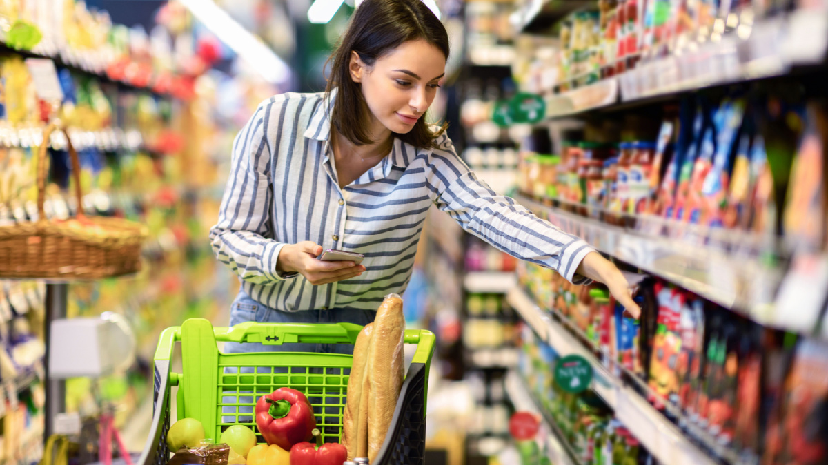 Portrait Of Millennial Lady Holding And Using Smartphone Buying Food Groceries Walking In Supermarket With Trolley Cart.