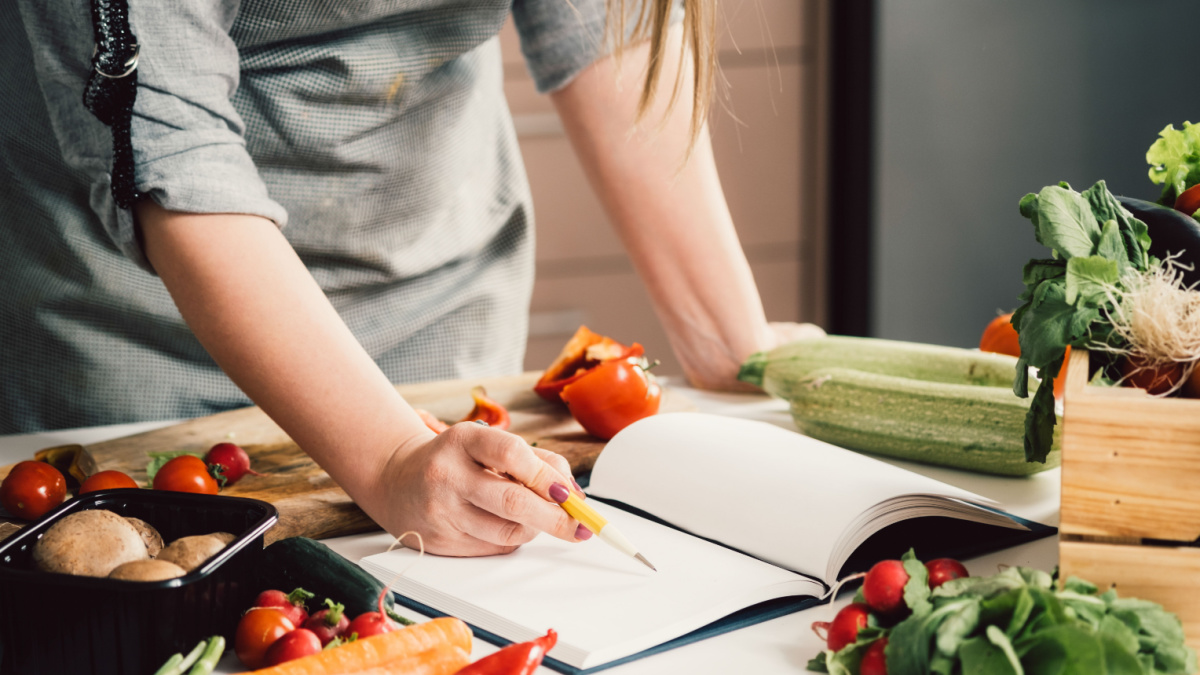 Unrecognizable woman is looking for recipes in cookbook. Female chef reading recipes in book and holding pen on table full with fresh vegetables for healthy lunch.