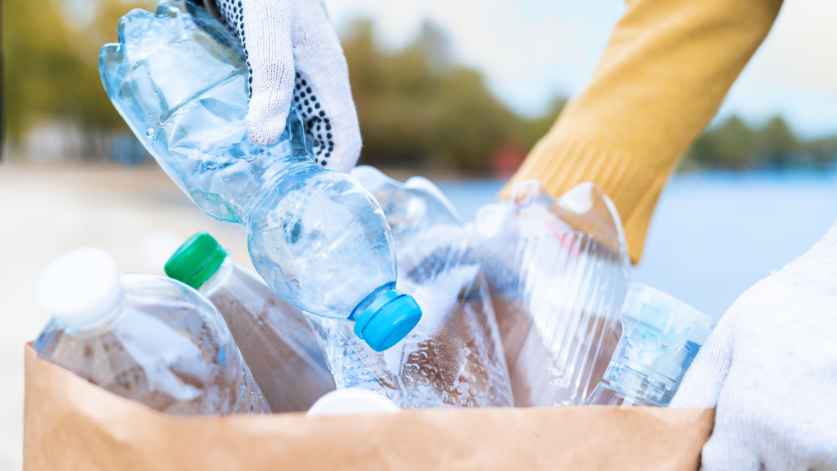 Young responsible female volunteer is collecting garbage on river ocean beach, folding plastic bottles in eco bag with recycle logo.