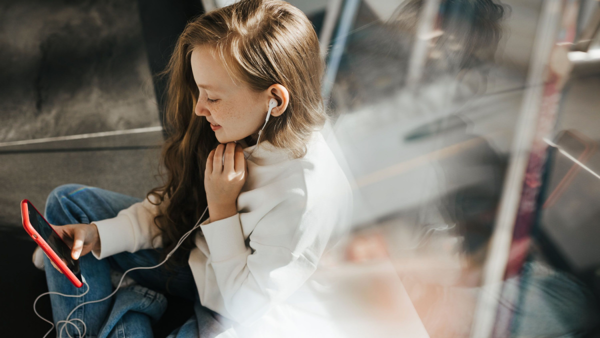 A teenage girl with blonde hair on her face with freckles is sitting on the floor in a shopping mall and looking at the phone and smiling, headphones in her ears.