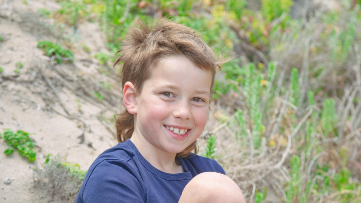 Aussie boy with mullet sitting on sand dune at the beach with big smile.