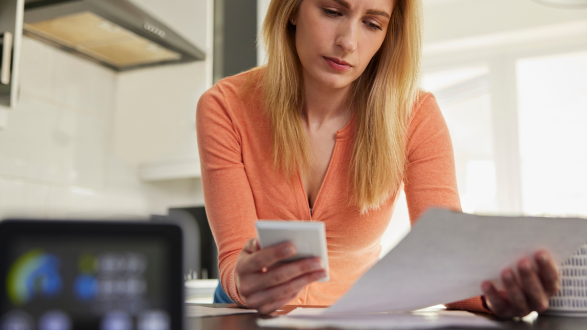 Smart Energy Meter In Kitchen Measuring Electricity And Gas Use With Woman Looking At Bills With Calculator.