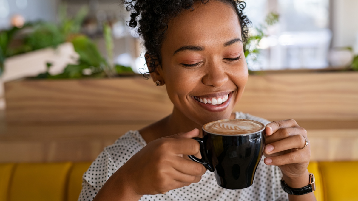 Smiling black young woman smelling freshly brewed coffee with eyes closed in cafeteria. Beautiful african girl smiling while relaxing in a coffee shop. Close up face of girl drinking latte coffee.