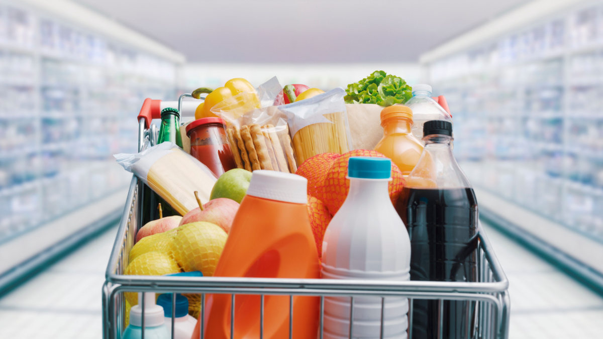 Shopping cart filled with food and drinks and supermarket shelves in the background, grocery shopping concept.