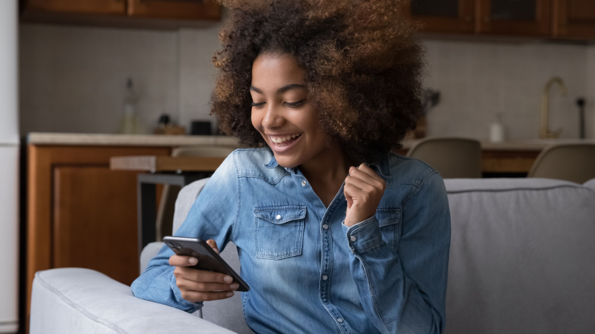 Excited cheerful gen Z African teenager girl using mobile phone, getting surprising good news, looking at screen, laughing, celebrating win, success, luck, achieve, good result.