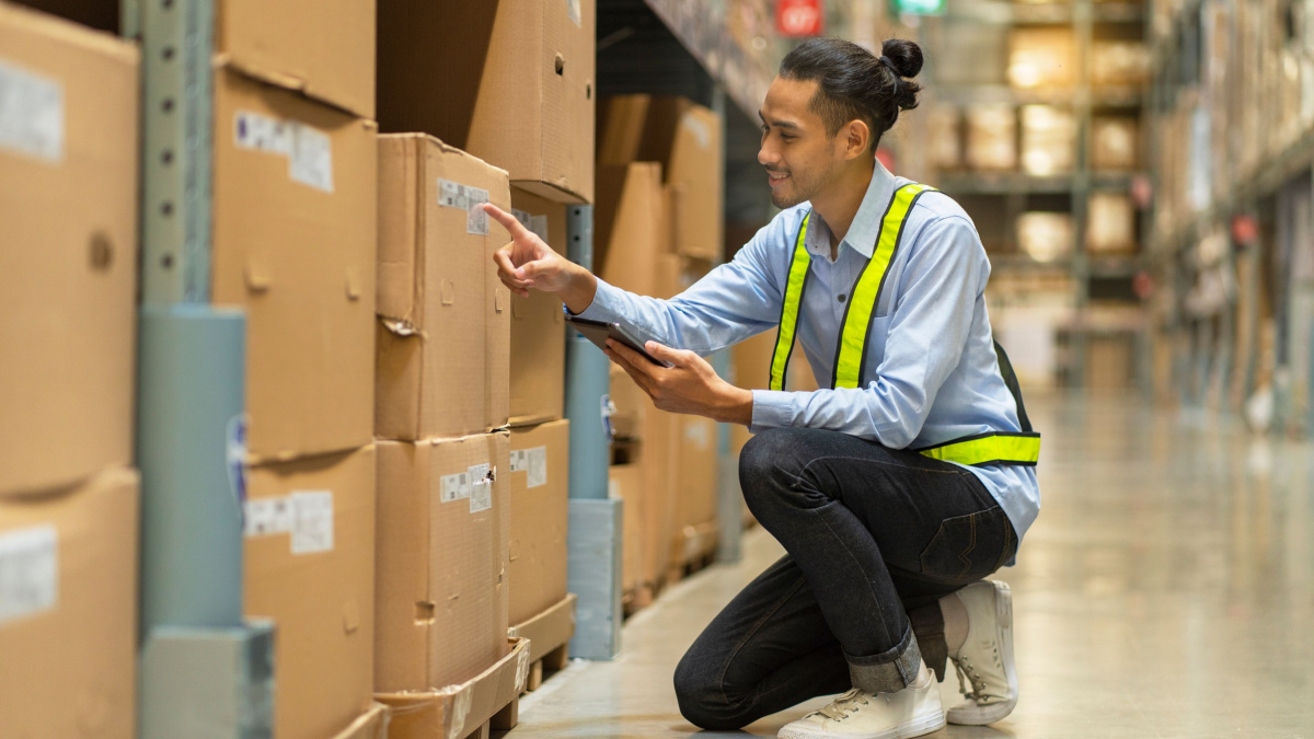 Asian man stocking worker holding tablet and checking inventory, counting the number of boxes at a wholesale store warehouse.