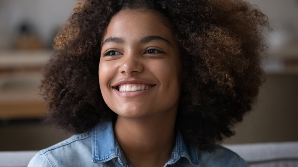 Close up beautiful African teenager girl sitting on sofa at home staring into distance, feel happy, having wide toothy charming smile and natural afro curly hairs posing indoors.