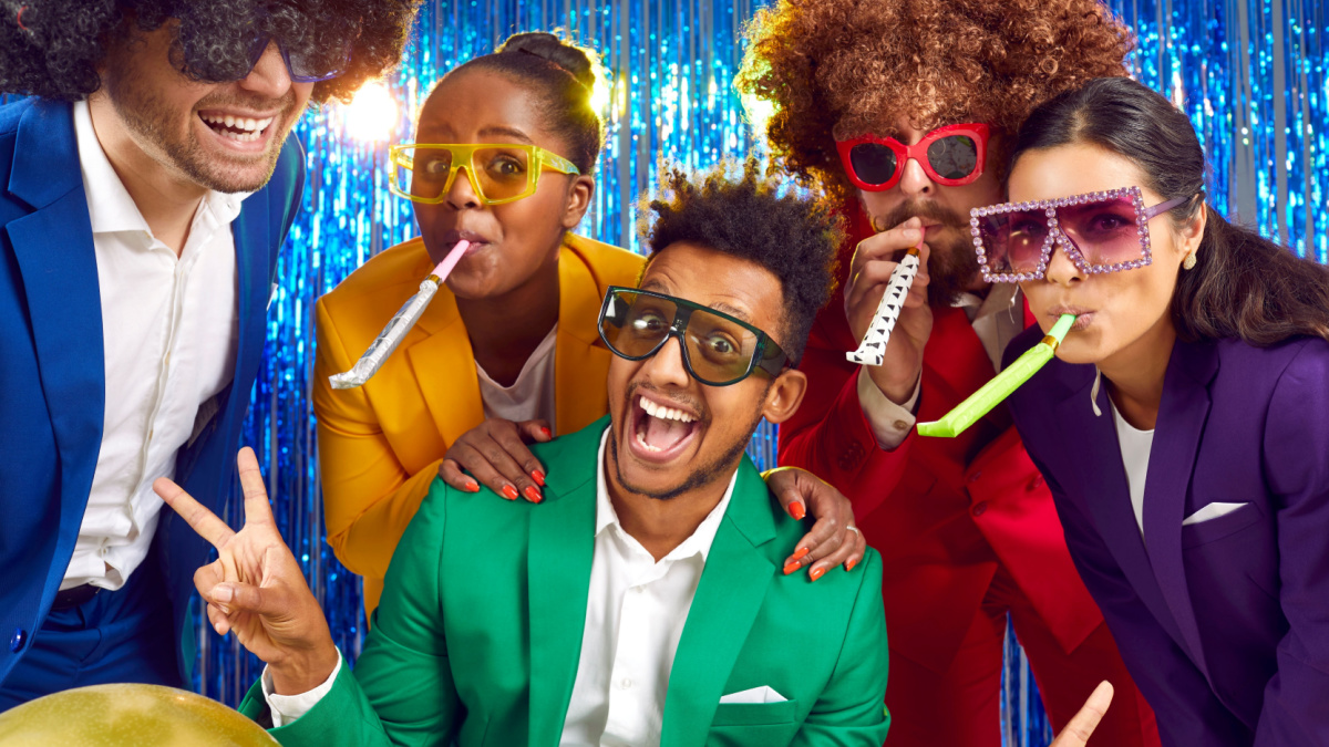Happy cheerful joyful smiling multiracial people wearing different bright yellow green red blue purple costumes, funny disco glasses, crazy curly wigs blow party horns on festive photobooth background.