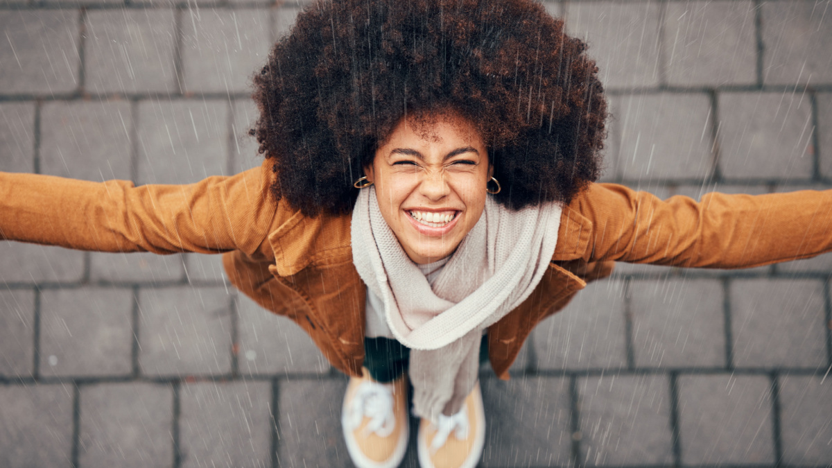 Black woman, stand to feel rain and happy in city in winter with smile, afro or trendy fashion.