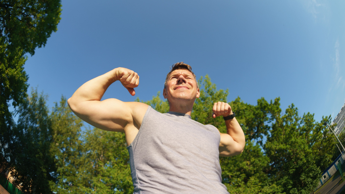 Handsome male model shows his muscles outdoors in sunny summer day. Male athlete is engaged on sports ground. He is funny. Concept of fitness, sport, strength exercising, training and lifestyle.