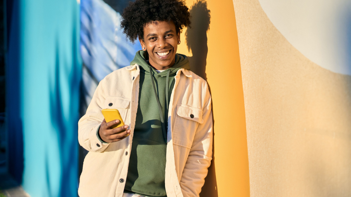 Smiling young cool African American guy holding mobile phone tech device standing at colorful city wall.