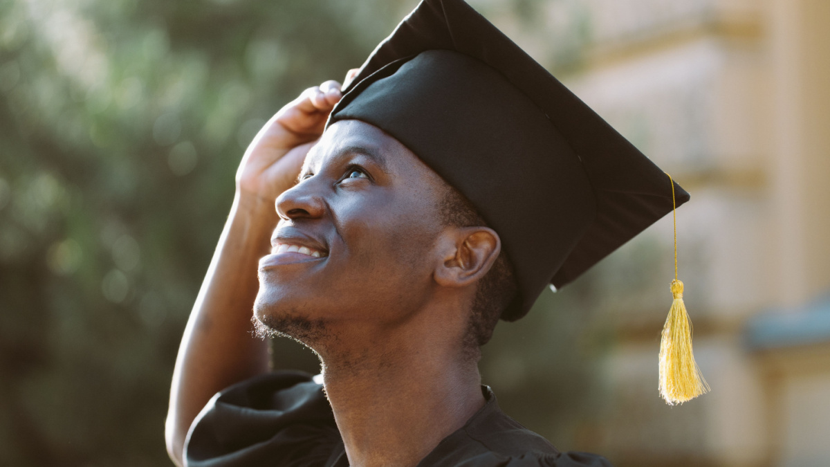 Portrait closeup of afro american student in graduation mantle and hat standing outdoors and with pleasure looking up. Job search, graduate from university, start in life, thoughts about future.