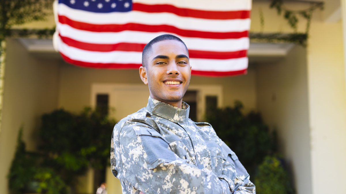 Portrait of happy biracial male american soldier wearing military uniform standing outside the house. American flag, patriotism and military service.