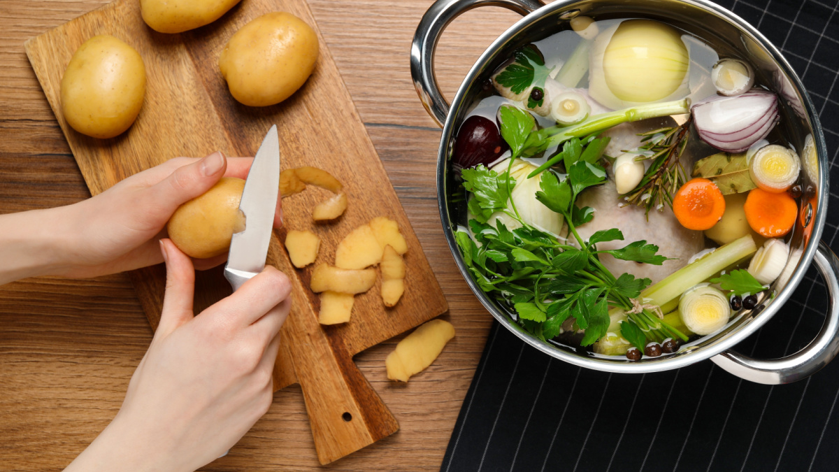 Woman peeling potato for cooking tasty bouillon at wooden table, top view.