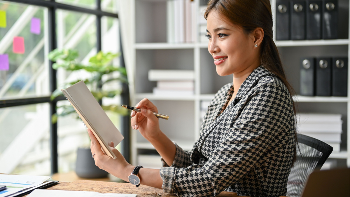 An attractive millennial Asian businesswoman or female boss makes an idea list in her notebook while sitting at her desk in the office.