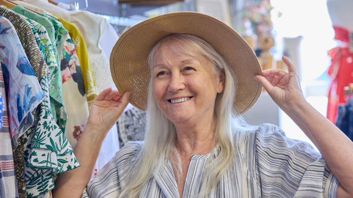 Senior Woman Buying Used Sustainable Clothes From Second Hand Charity Shop Or Thrift Store Trying On Hat.