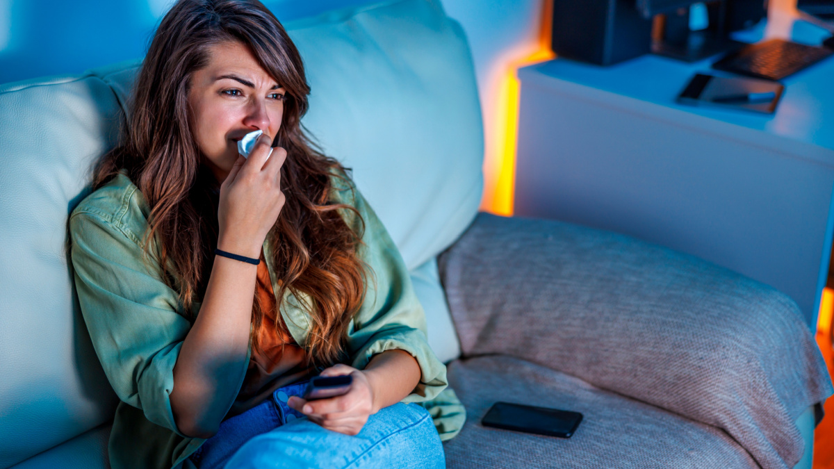 Beautiful young woman sitting on couch late at night, sad while watching drama movie on TV, crying and wiping tears with paper tissues.