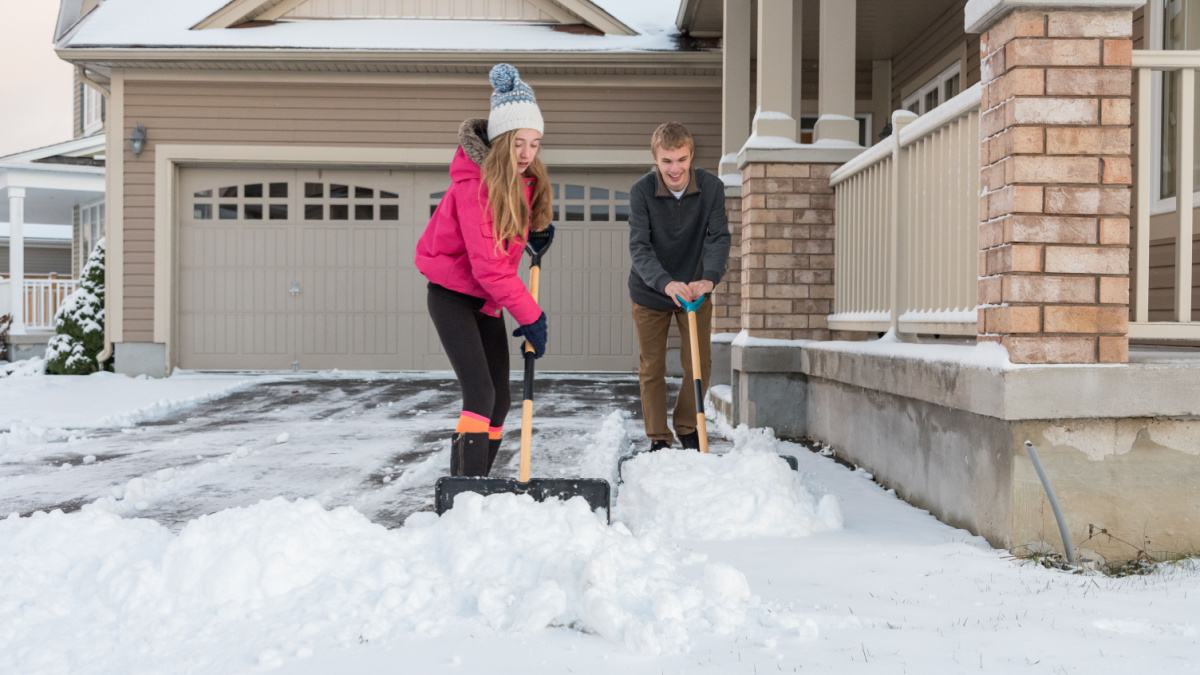 A teenage girl and boy shoveling a snow covered suburban house driveway during winter.