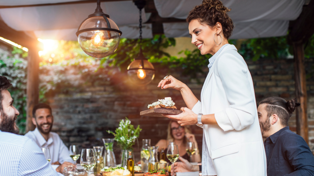 Beautiful Caucasian smiling woman putting a plate with meal on dining table at backyard celebration.