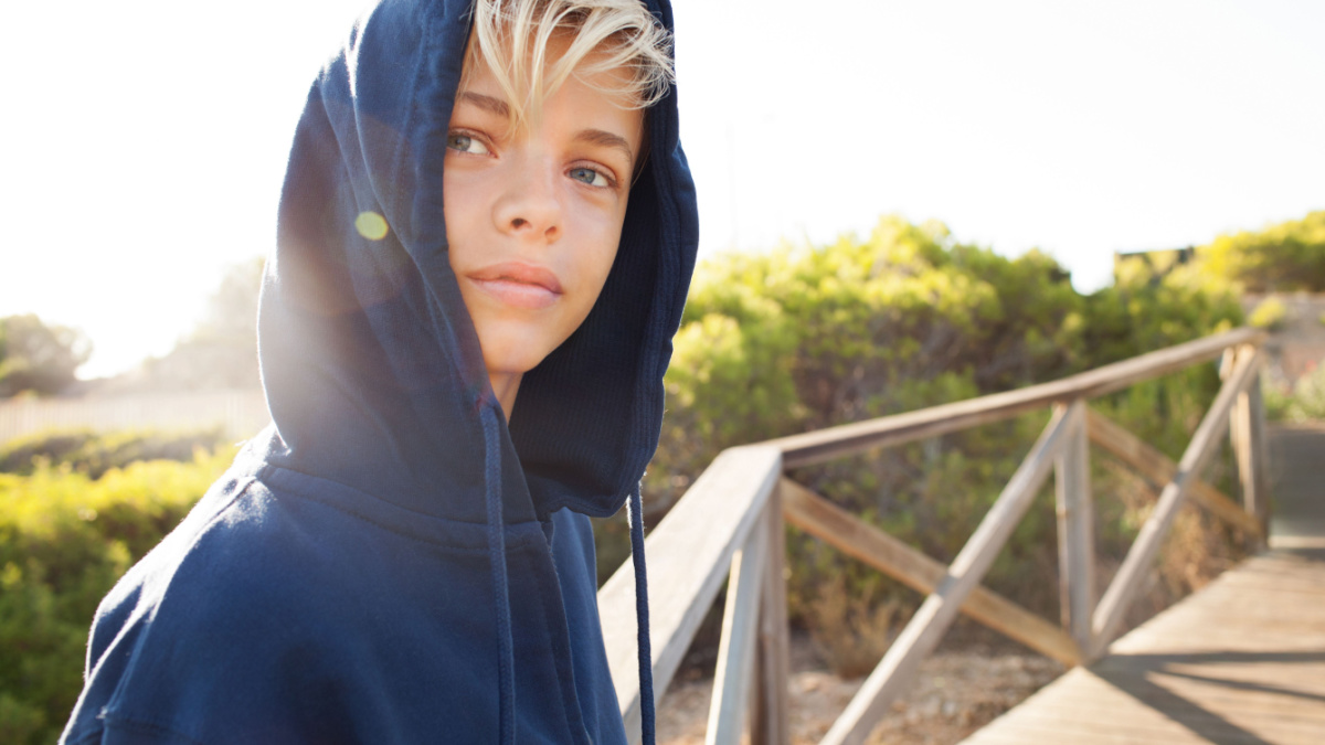 Portrait of beautiful young man wearing hoody in nature park with sun flare, serene smiling looking away, leaning on banister, outdoors.