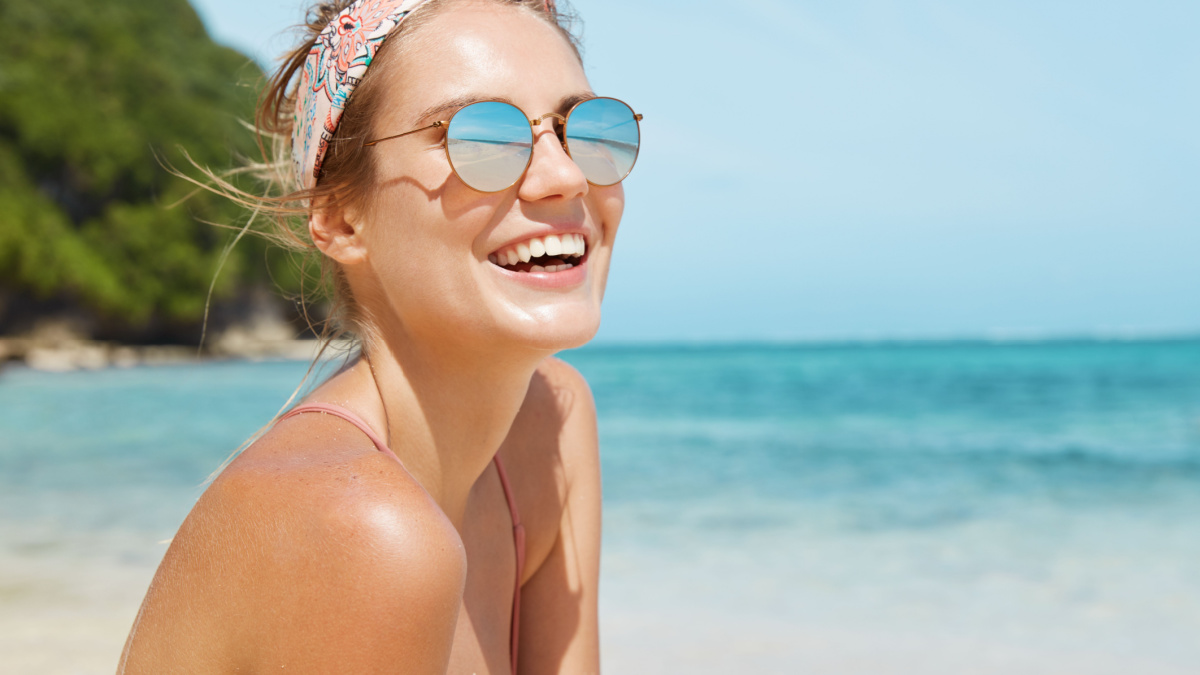 Close up shot of pleasant looking female wears headband and sunglasses, travels in tropical country, poses against warm peaceful ocean, blue sky and rocky cliff in background. Good relaxation