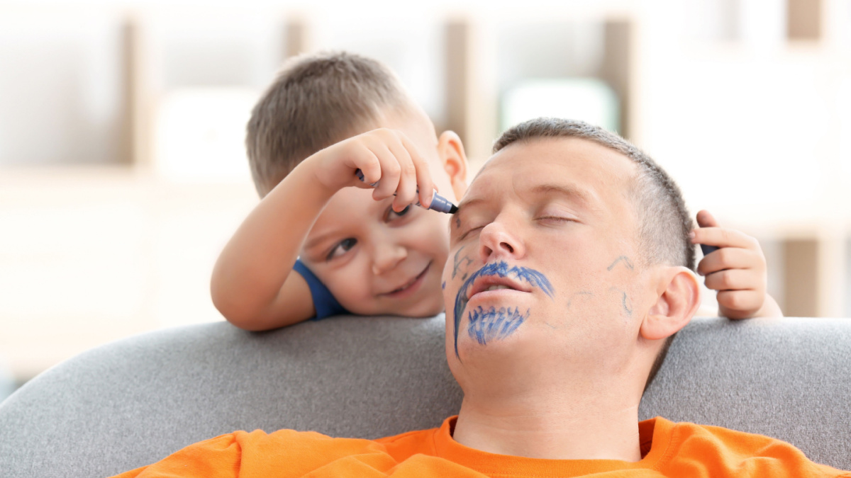 Little boy painting his father's face while he sleeping. April fool's day prank.