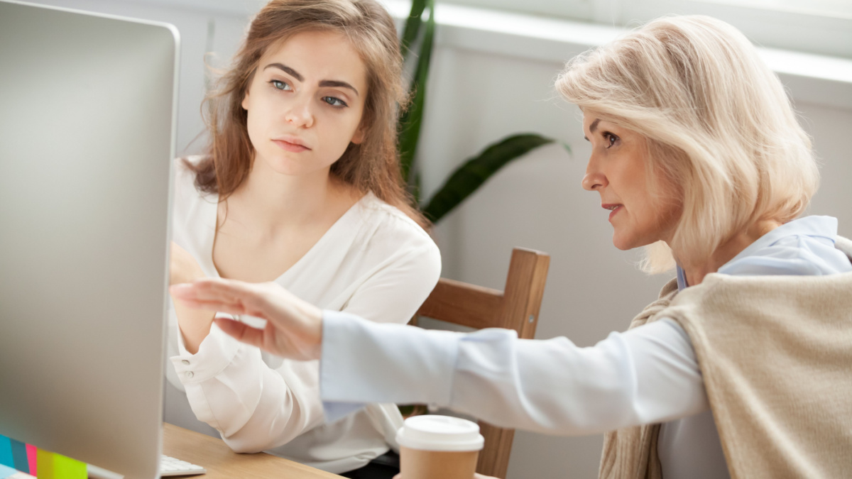 Senior and young female colleagues discuss online project look at pc screen, older mentor teaches young woman explains corporate software work, aged executive helps intern, teamwork on computer task.