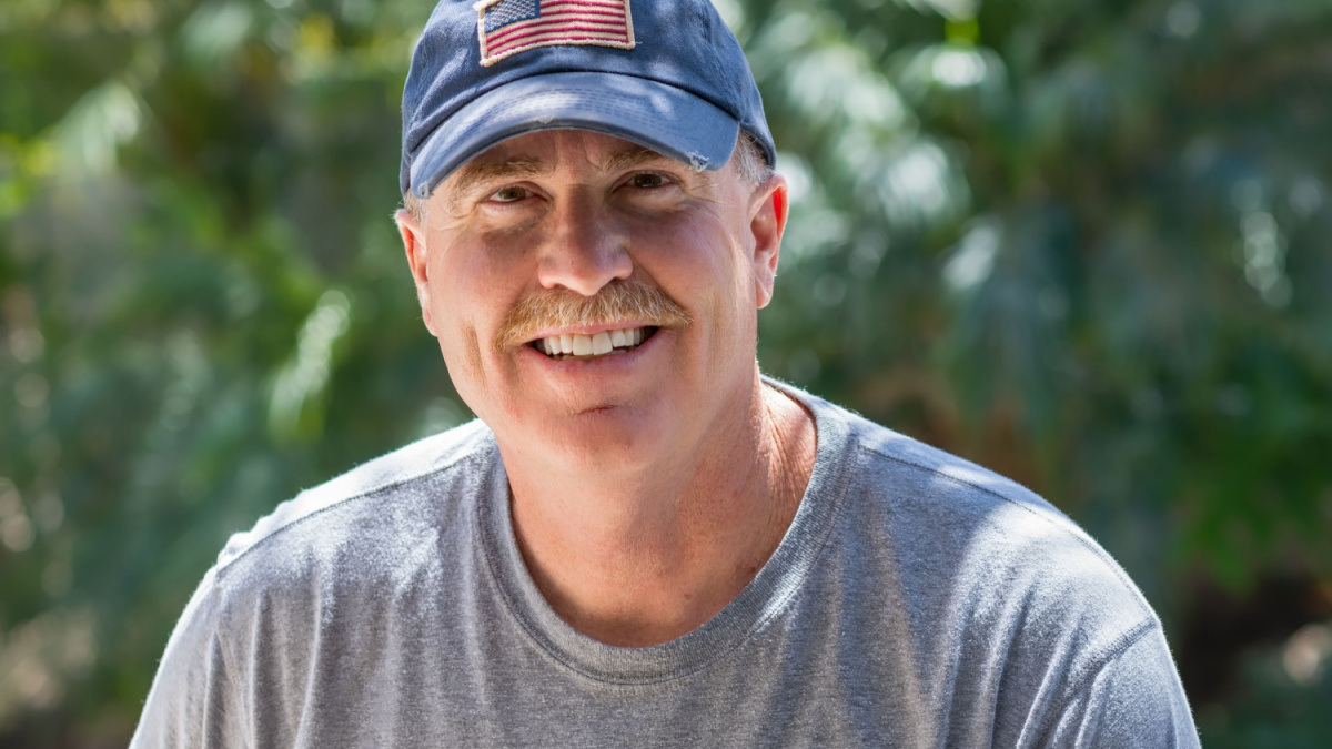 Older male smiles warmly outside wearing USA flag baseball hat.