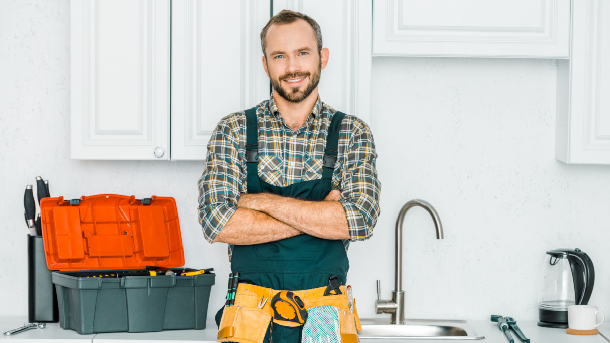 smiling handsome plumber standing with crossed arms and looking at camera in kitchen