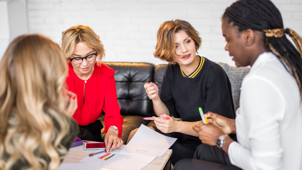 Group of international MLM partners discuss their work in co-working space talking and smiling while sitting at the desk table with smartphones and drawings on paper.