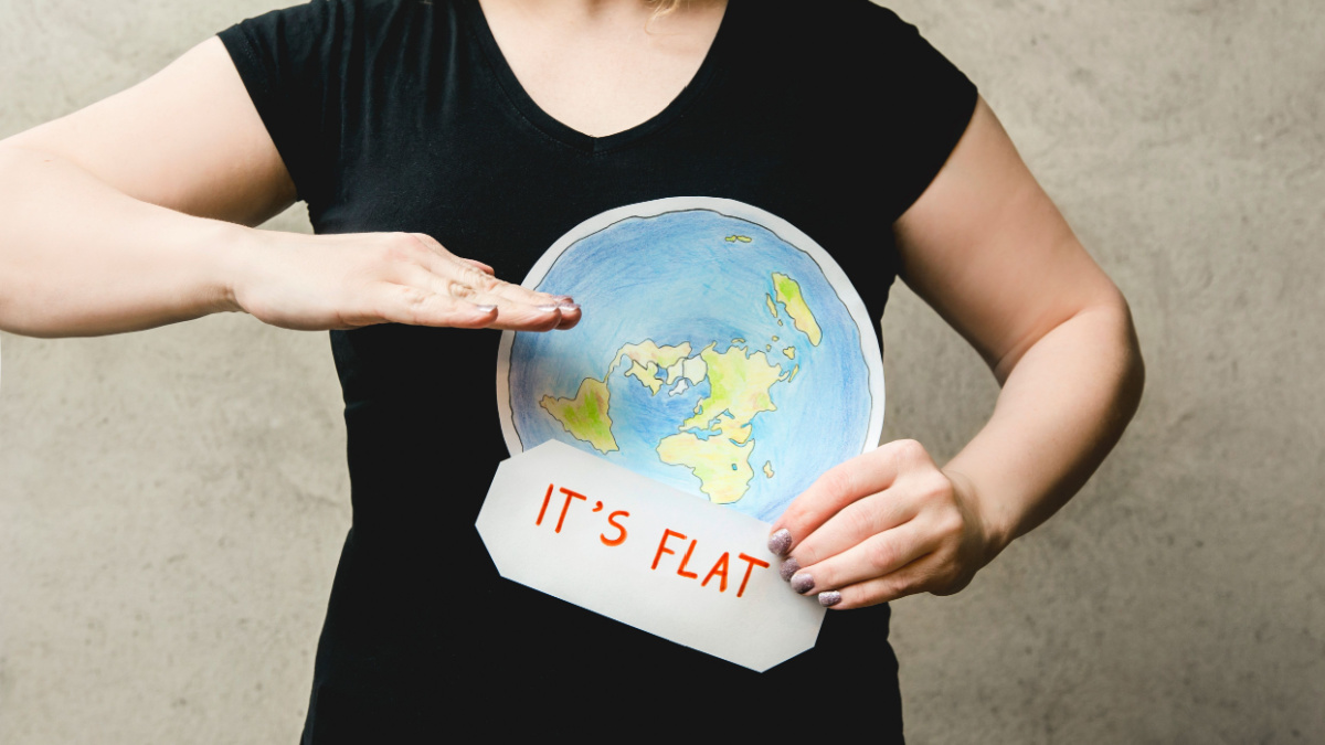 Flat Earther concept. Person who believes that Earth is flat disc. Anonymous woman holding flat Earth model in front of body with text: It`s flat. Isolated on gray background, studio shot.