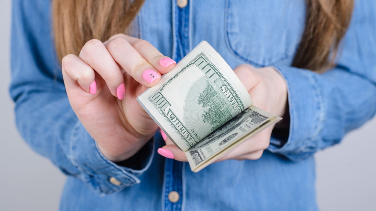 Cropped close-up photo of girlish beautiful hands with long nails pink bright manicure on fingers holding calculating old money wearing denim clothes isolated grey background.