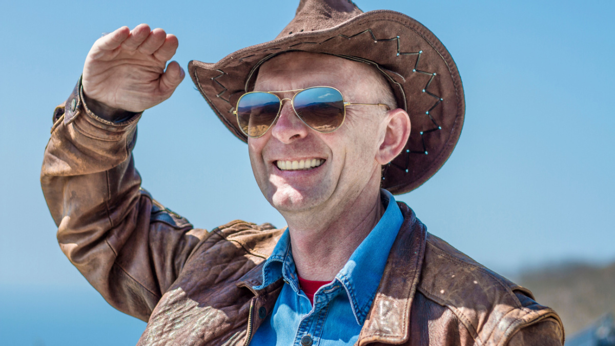 In the mountains there is a man in a cowboy hat, leather jacket, glasses. A man puts his hand to his hat. Background of mountains and sky.