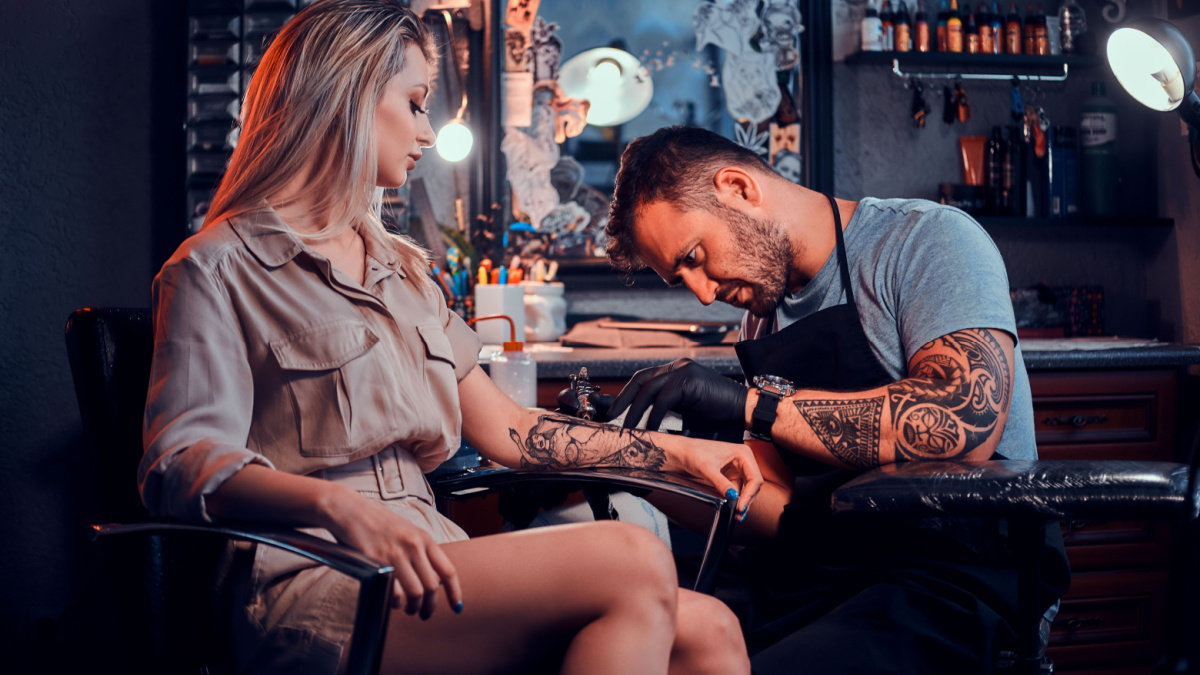 Dilligent focused tattoo artist is creating new tattoo on young woman's hand at tatoo studio.