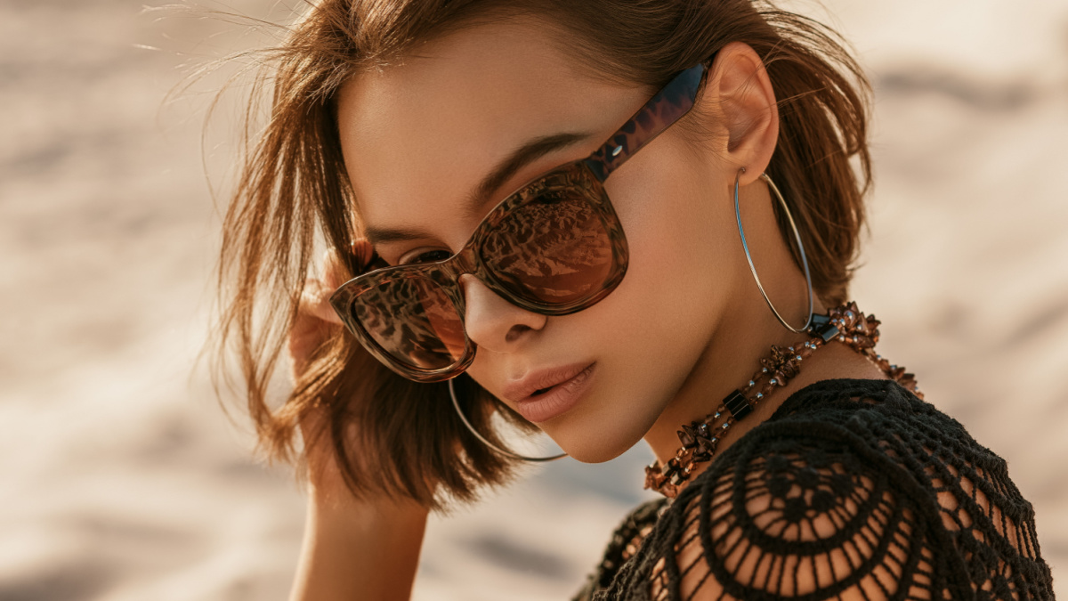 Outdoor close up portrait of young beautiful woman with tanned skin wearing luxury wide frame square sunglasses, hoop earrings, crochet top, posing on sand, at sunset.