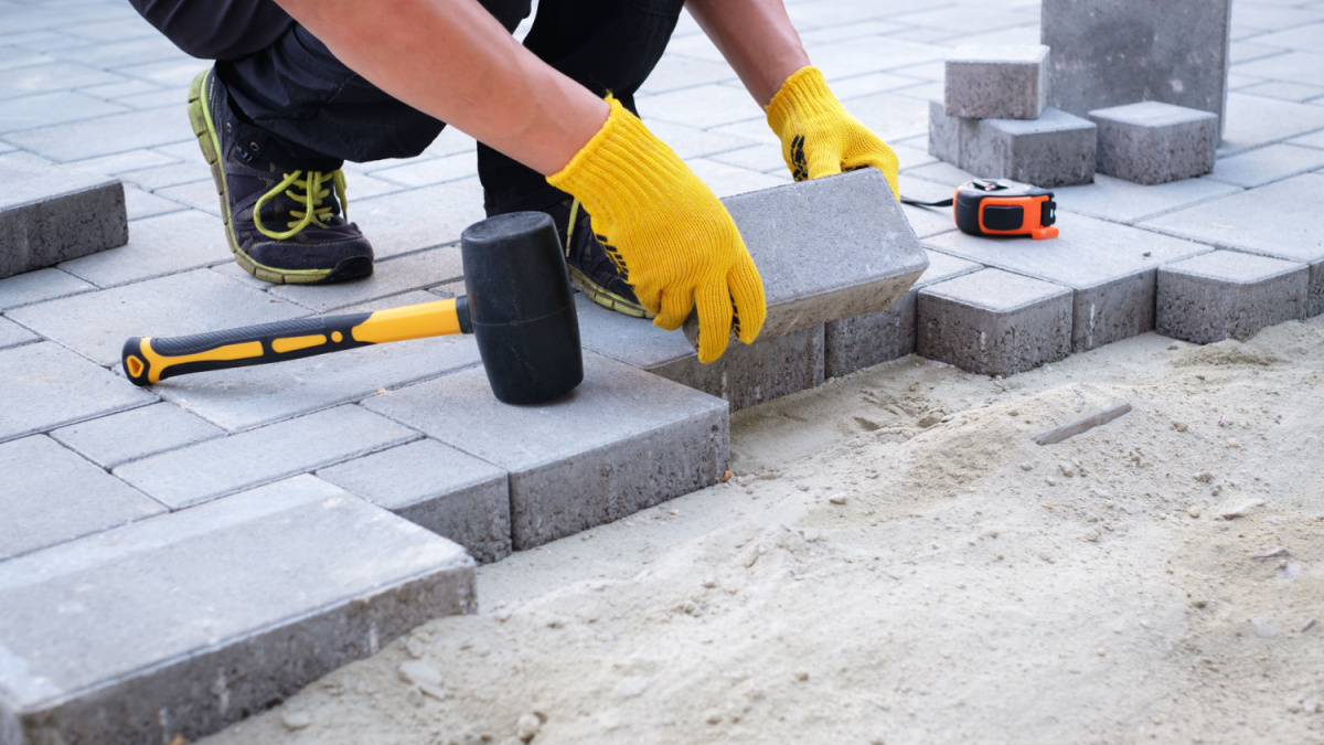 The master in yellow gloves lays paving stones in layers. Garden brick pathway paving by professional paver worker. Laying gray concrete paving slabs in house courtyard on sand foundation base.