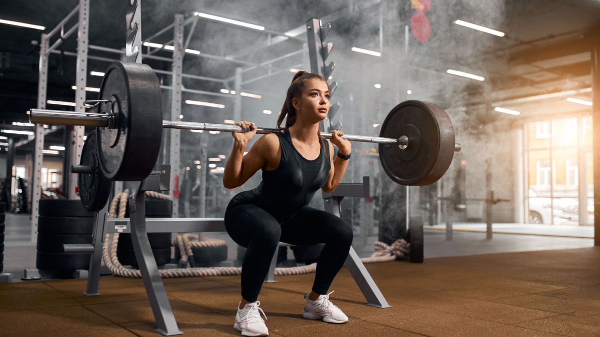 Beautiful young powerlifter squatting in modern fitness studio using heavy barbell, looking aside expressing self assurance, professional sport concept, white smoke in the air