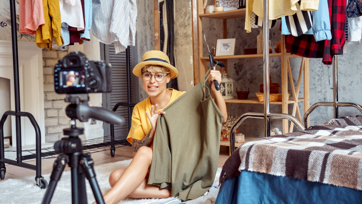 Young woman fashion blogger influencer wearing hat and eyeglasses sitting on fluffy carpet at urban stylish apartment recording video vlog on digital camera telling clothing tips positive